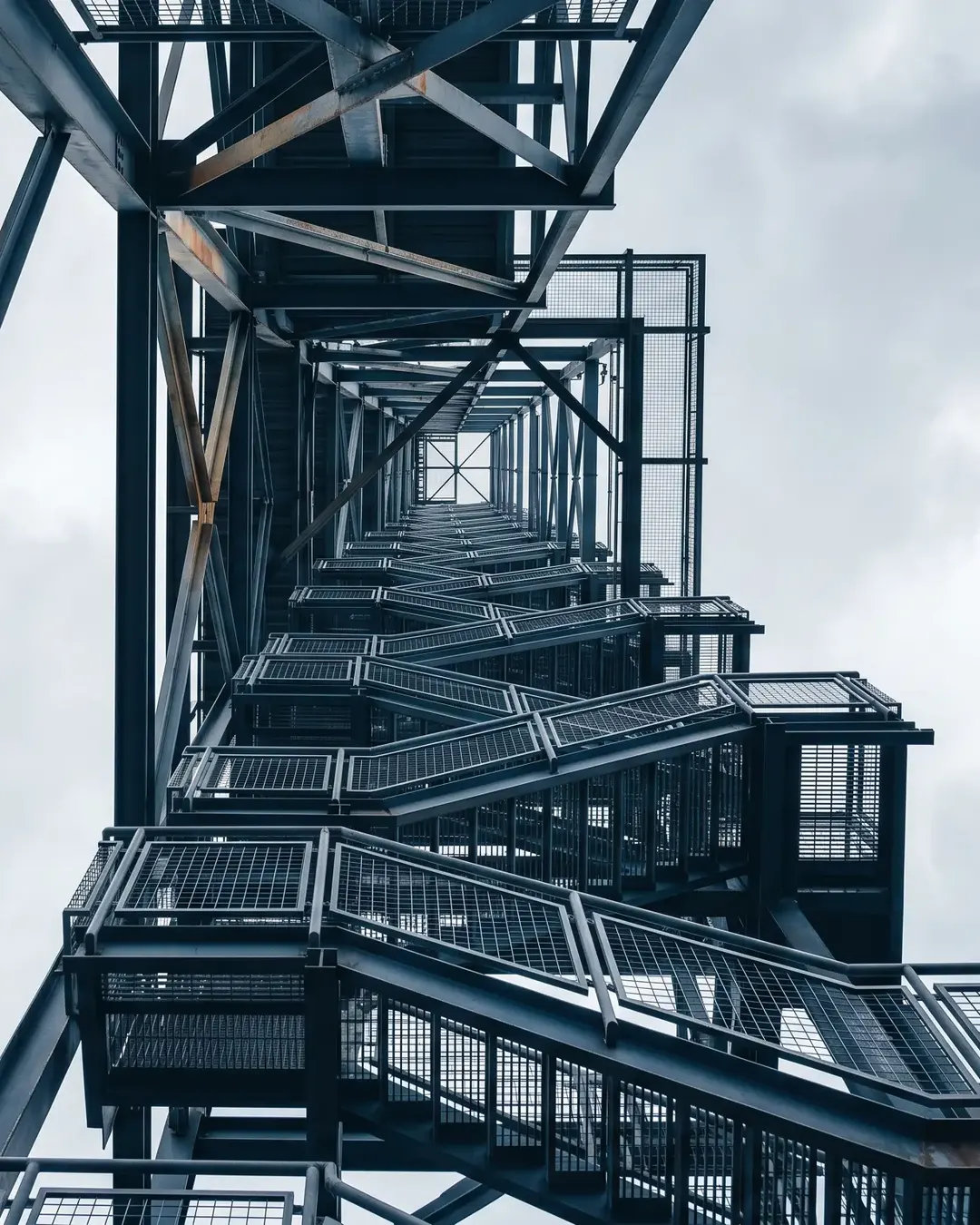 A steel access gantry and stair tower against an overcast sky, photographed from directly beneath, empty of people