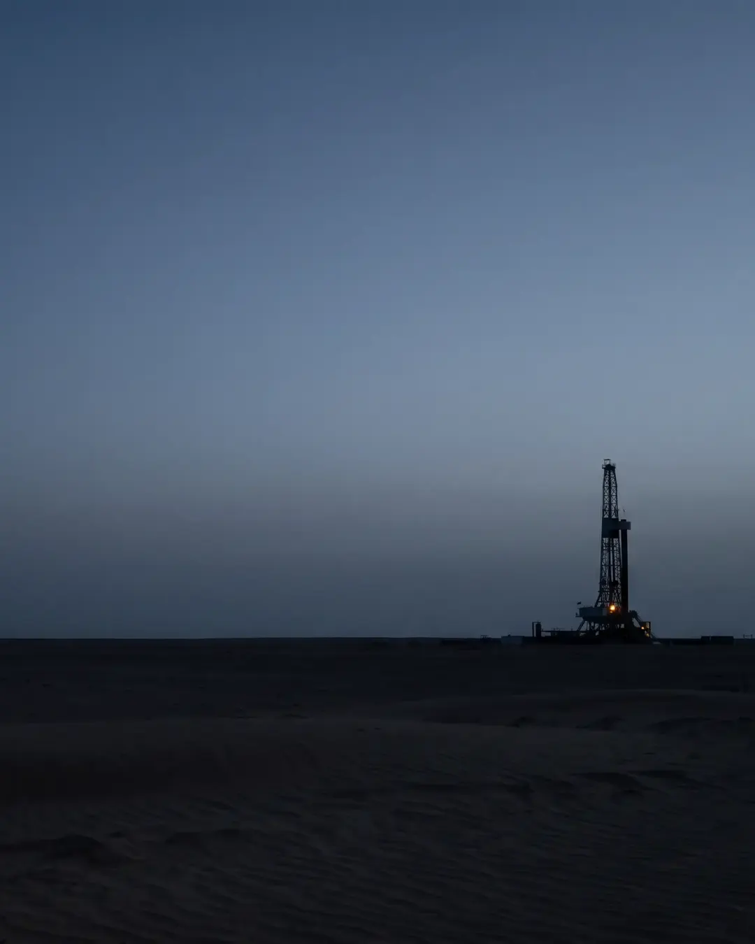 A single drilling derrick silhouetted at the edge of a wide desert horizon at dusk
