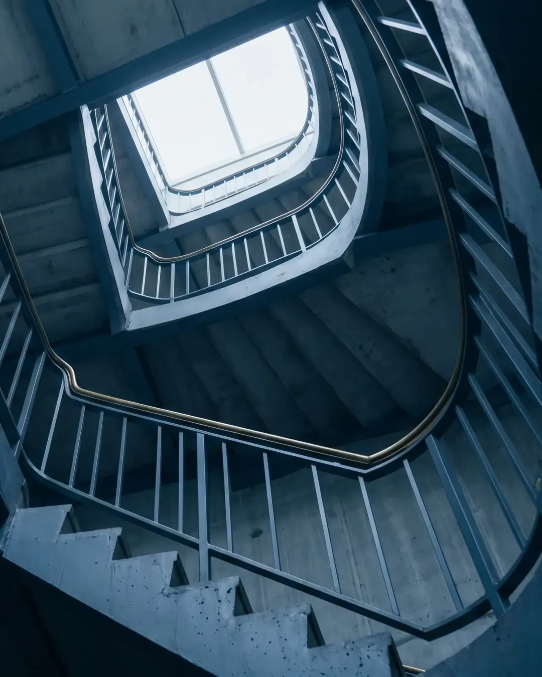 A cold-lit concrete stairwell photographed looking upward through five flights toward a skylight
