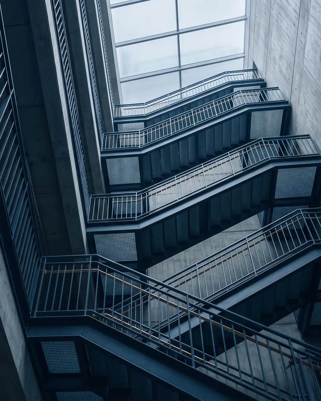 An industrial stairwell rising through five flights of exposed steel and board-marked concrete toward an overcast skylight